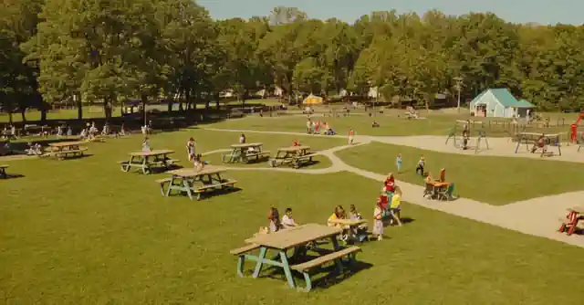 People at an open field under the sun with picnic tables and childrens playground equipment.