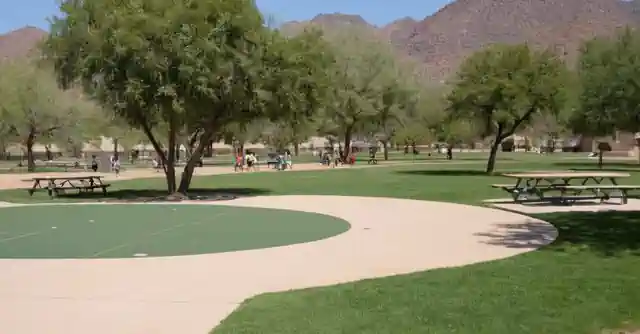 Image showing various facilities like sports fields, picnic areas at Indian Bend Park in Phoenix. Note that this instruction assumes some knowledge about the facilities and location which is assumed to be available within the described image itself without needing explicit clarification about it (a park map would also help). Thus, this could involve:- Depictions of the different sports fields - grassy with goals or baskets as required for soccer, basketball etc. These facilities should appear open, in use and busy to reflect their availability. - Picnic areas that can be represented by people having picnics, families setting up a barbeque or playing outdoor games on the well-prepared ground covered with grass and shady trees. The benches could also help indicate its purpose for relaxation and dining area.- A signboard indicating the Indian Bend Park name can enhance comprehension of context along with other services like restroom facilities, water fountains etc to depict that it isnt just limited to recreational activities but involves several community service aspects as well. Remember, for making any public place image more relatable and attractive, the presence of families with children (in a safe distance due to social distancing policies, if applicable) can be included which will also convey that its a family-friendly spot.