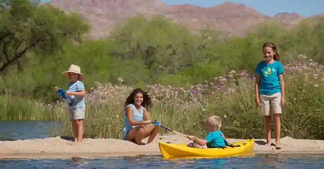 Children happily participating in various activities by the lake. Short captions on their joy and learning being tagged along, emphasizing ideal for children of Scottsdale. at or near each activity - a clear sky with swallows flying over (bird watching), kids building sandcastles (sandcastle building activity), colorful wildflowers blooming (botany exploration) and boats floating gently on the waters (kayaking instruction).