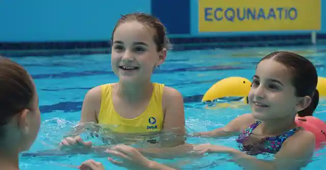 A swimming facility sign above a crystal blue pool with children learning to swim.