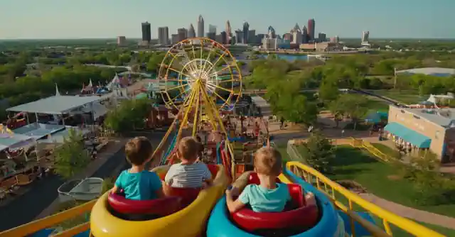Children playing on amusement rides in a theme park with the Peoria skyline in the background.
